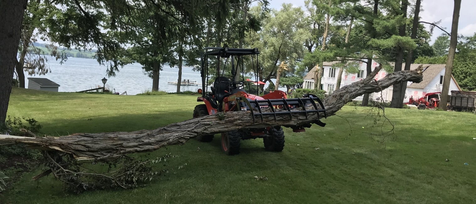 Red Kubota tractor moving a large log with a grapple on a lakeside lawn